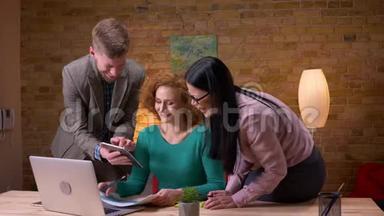 三名办公室工作人员用笔记本电脑平板电脑和图表讨论数据的特写镜头，在室内开心地笑着